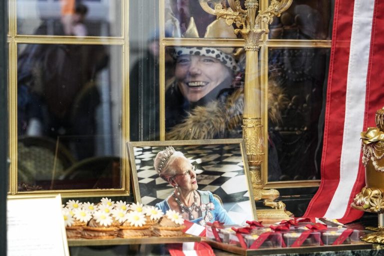 A woman wearing a crown smiles behind a window of a cafe where a picture of Danish Queen Margrethe is on display in Copenhagen, Denmark, Sunday, Jan. 14, 2024. Denmark's Crown Prince Frederik takes over the crown on Sunday from his mother, Queen Margrethe II, who is breaking with centuries of Danish royal tradition and retiring after a 52-year reign. (AP Photo/Martin Meissner)