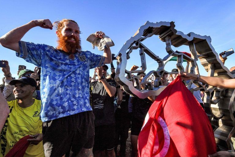 British runner Russ Cook gestures as he poses for a picture with the memorial sign marking the northern-most point of Africa upon arrival at Cape Angela, northeast of Tunis, on April 7, 2024 while surrounded by supporters who joined him for the final leg of the 16,000 kilometre challenge to cross the African continent from South Africa's Cape Agulhas to Tunisia's Cape Angela to raise money for charity. (Photo by FETHI BELAID / AFP)