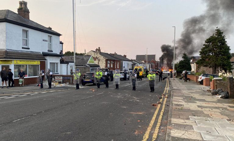Smoke billows from a fire started by protesters as riot police stand guard after disturbances near the Southport Islamic Society Mosque in Southport, northwest England, on July 30, 2024, a day after a deadly child knife attack. Violent clashes broke out in the northern England town where a knife attack claimed the lives of three children, with around 100 protesters lighting fires and battling police. A 17-year-old male suspect from a nearby village arrested shortly after the incident remained in custody, police added, as they warned against speculating about his identity or details of the investigation. (Photo by Roland LLOYD PARRY / AFP)