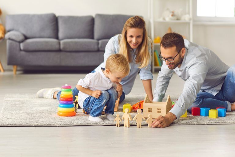 Young parents play with their son wooden figurines sitting on the floor among various toys. Mother, father and child spend time at home in a cozy children's room. Family holiday concept.